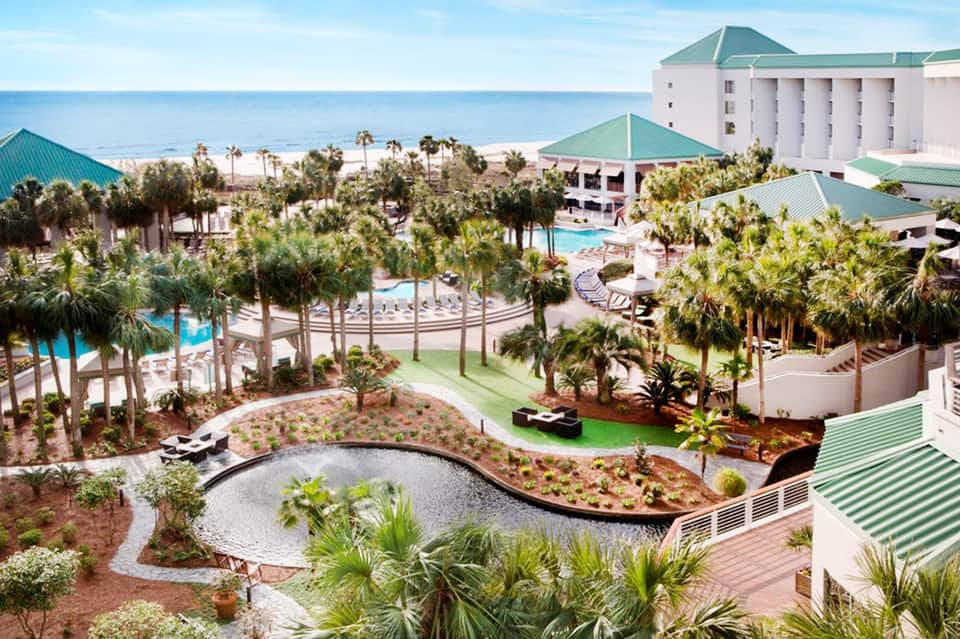Aerial view of resort with multiple pools, palm trees, and white buildings with green roofs near beach