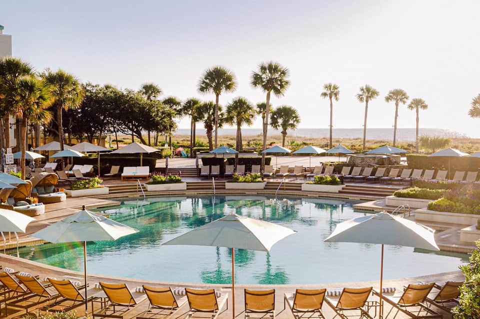Expansive resort pool with white umbrellas, palm trees, and beachfront view
