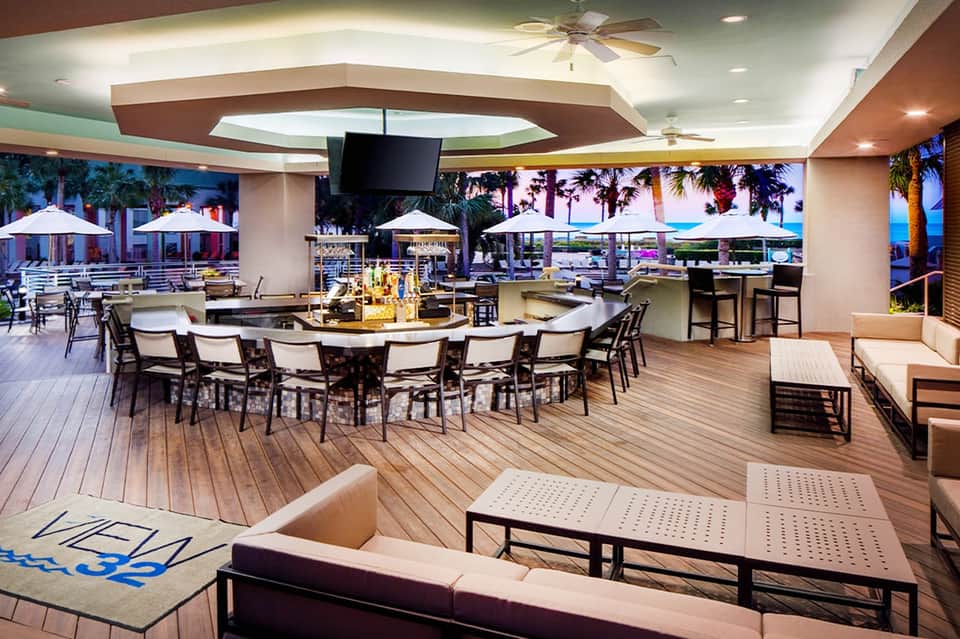 Seaside terrace lounge with bar, dining areas, white umbrellas, palm trees, and ocean views