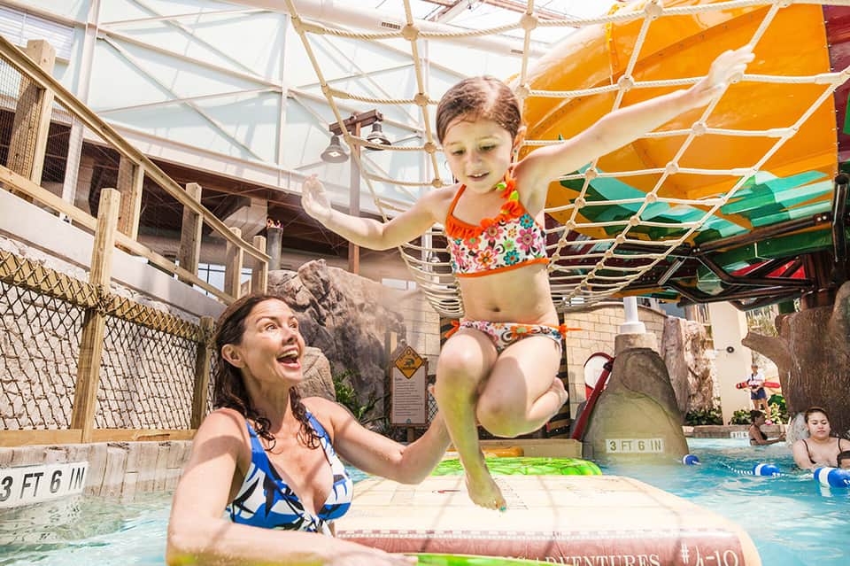 Child jumping into indoor pool while woman watches, under colorful sail canopy