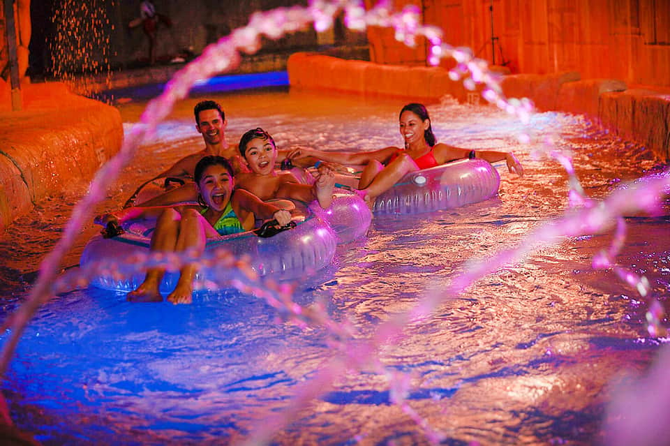 Family playing in glowing purple-lit indoor pool with illuminated floats and inflatable toys