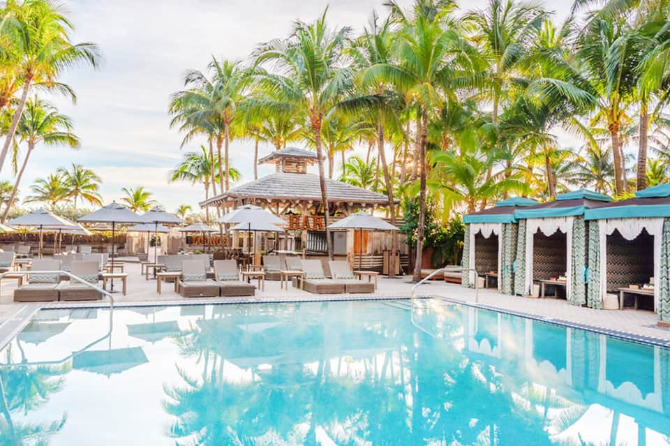 Luxurious resort pool surrounded by palm trees, lounge chairs with umbrellas, cabanas, and tropical architecture