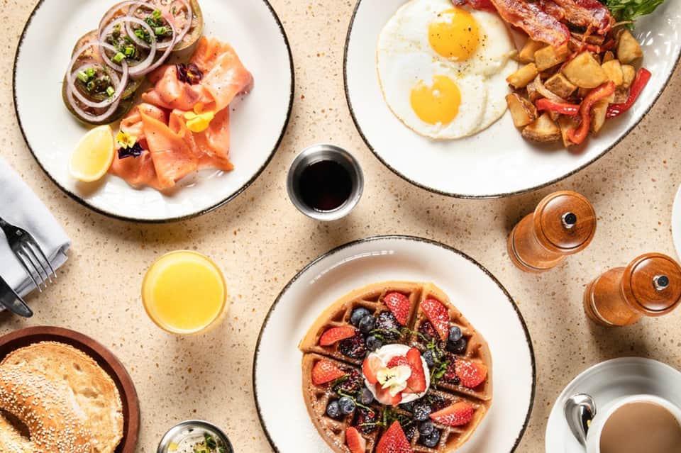 Overhead view of breakfast spread featuring waffles, eggs, bacon, salmon, and fresh fruit on white plates