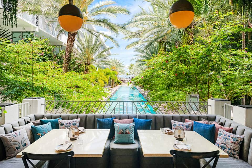 Upscale poolside dining area with palm trees, hanging orange lights, and blue cushioned seating