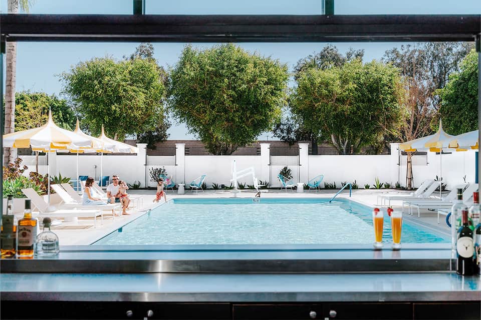 Indoor-outdoor pool view with lounge chairs, white umbrellas, trees, and swimmers visible from above