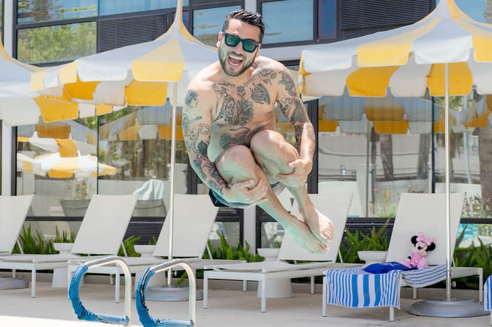 Tattooed man holding child by pool with yellow and white striped umbrellas in background
