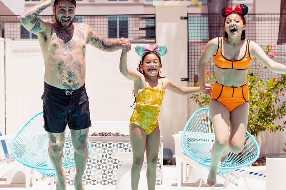 Family of three celebrating in pool wearing Mickey ears and colorful swimwear on rooftop deck