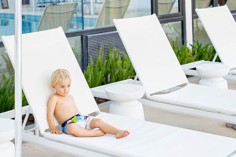Young child sitting in white lounge chair by resort pool and building