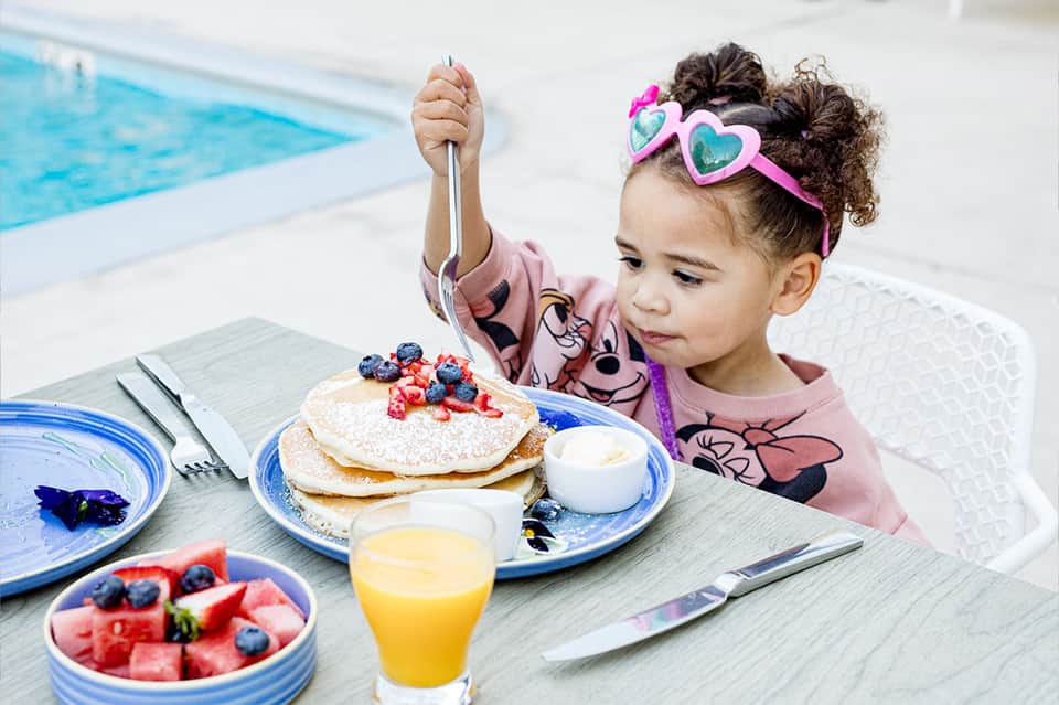 Young child enjoying pancakes and fruit by the pool with juice and syrup