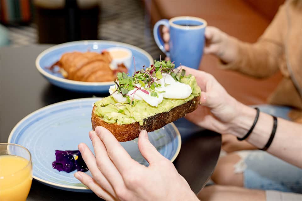 Person holding avocado toast with microgreens and blue coffee mug at breakfast