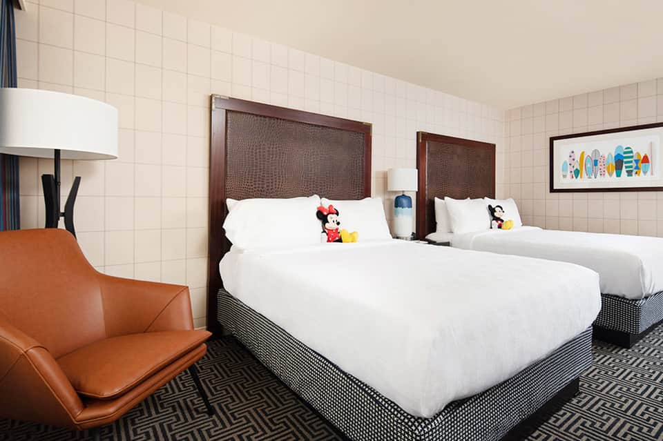 Hotel room with queen beds, dark wood headboards, white linens, orange accent chair, and Disney character pillows