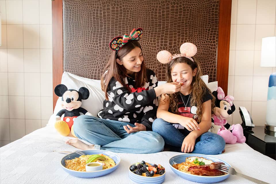 Two children in Disney-themed room with character plushies, breakfast plates, and decorative headbands