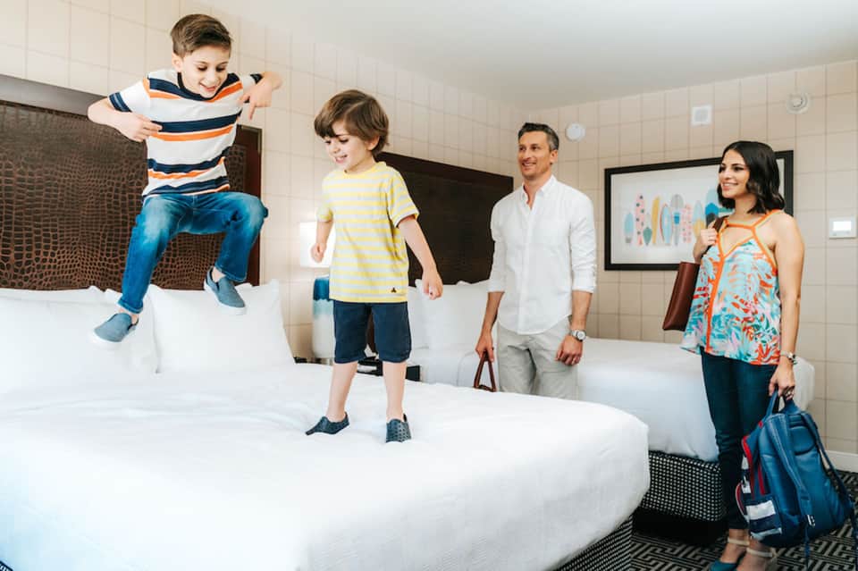 Family in modern hotel room with children jumping on beds while parents watch with luggage nearby
