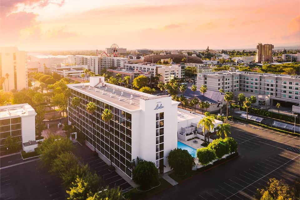 Aerial view of modern hotel with pool at golden hour, surrounded by palm trees and residential area
