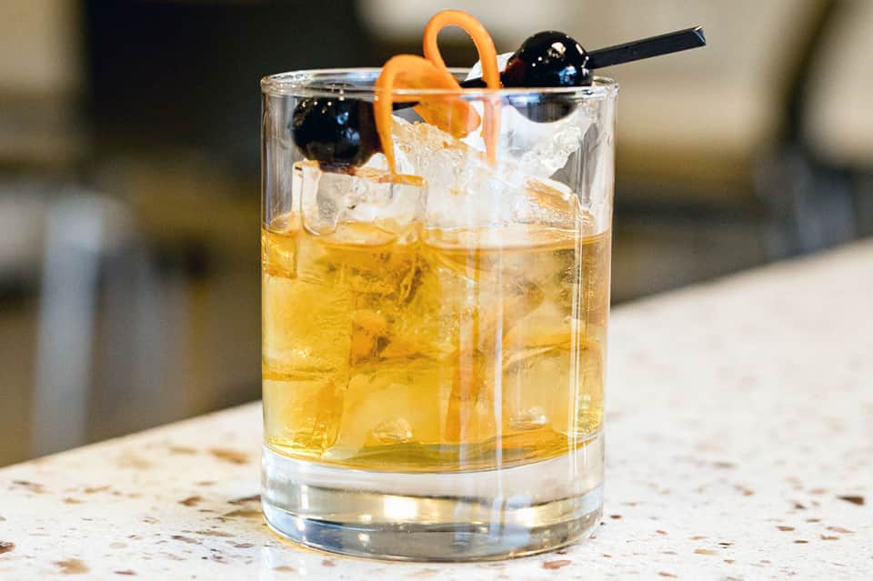 Golden cocktail with ice, black olives, and orange peel garnish on stone countertop