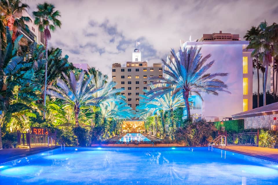 Illuminated resort pool at dusk surrounded by palm trees and colorful buildings