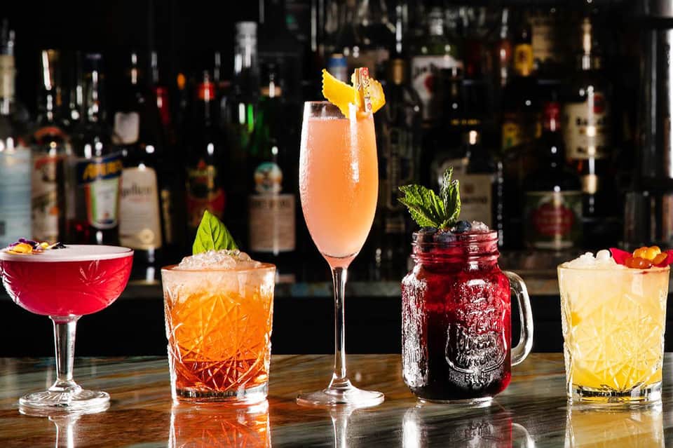 Array of five colorful cocktails at bar with bottle collection background