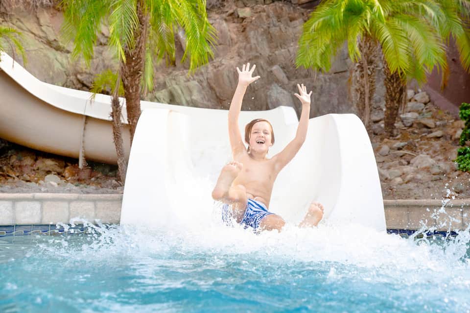 Child sliding down white water slide into pool with palm trees and rocky backdrop
