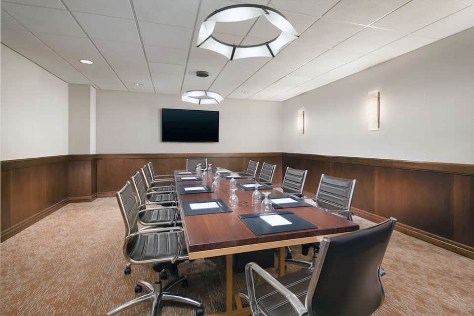 Hotel conference room with long wooden table, office chairs, TV screen, and wood paneling