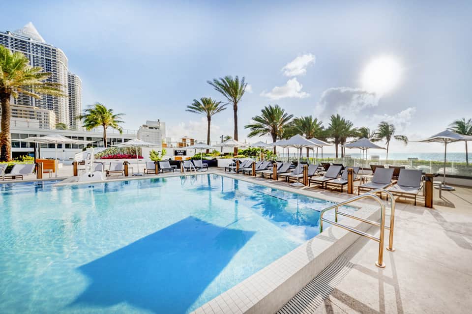 Rooftop outdoor pool with turquoise water, palm trees, lounge chairs, umbrellas, and high-rise building backdrop.