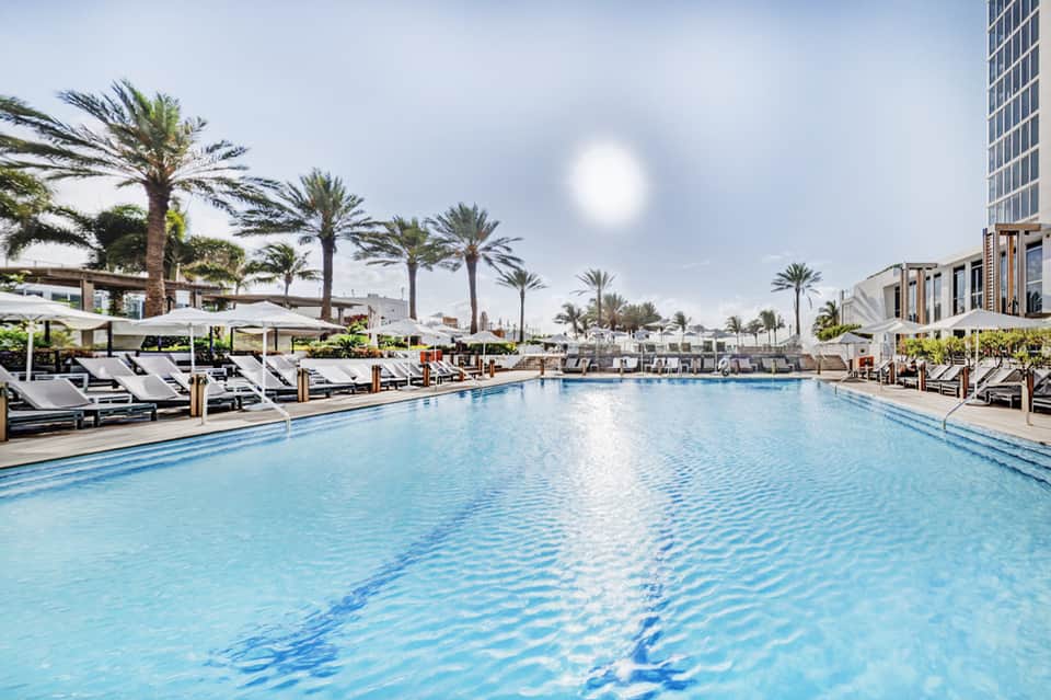 Large resort pool with palm trees, white lounge chairs, and modern building architecture