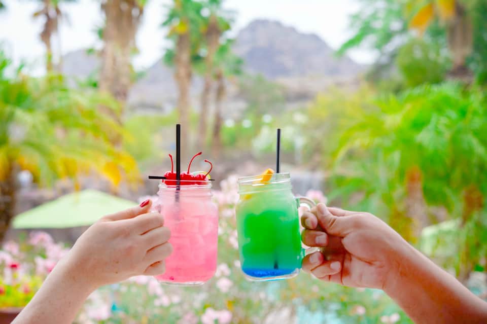 Two colorful tropical cocktails with straws held outdoors with desert mountains and palm trees in background