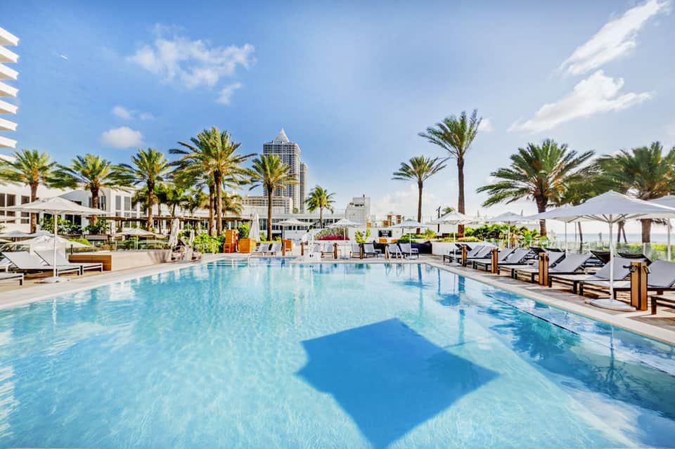 Resort swimming pool with palm trees, white umbrellas, lounge chairs, and city skyline