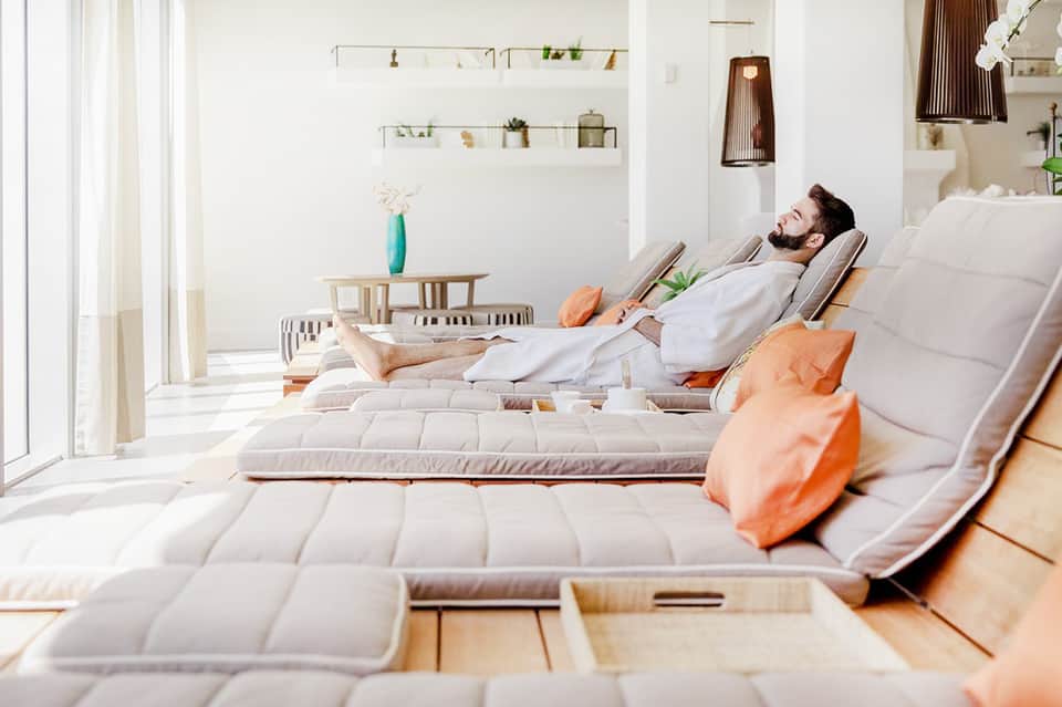 Man relaxing on white spa lounger with orange accent pillows in minimalist wellness room with natural light