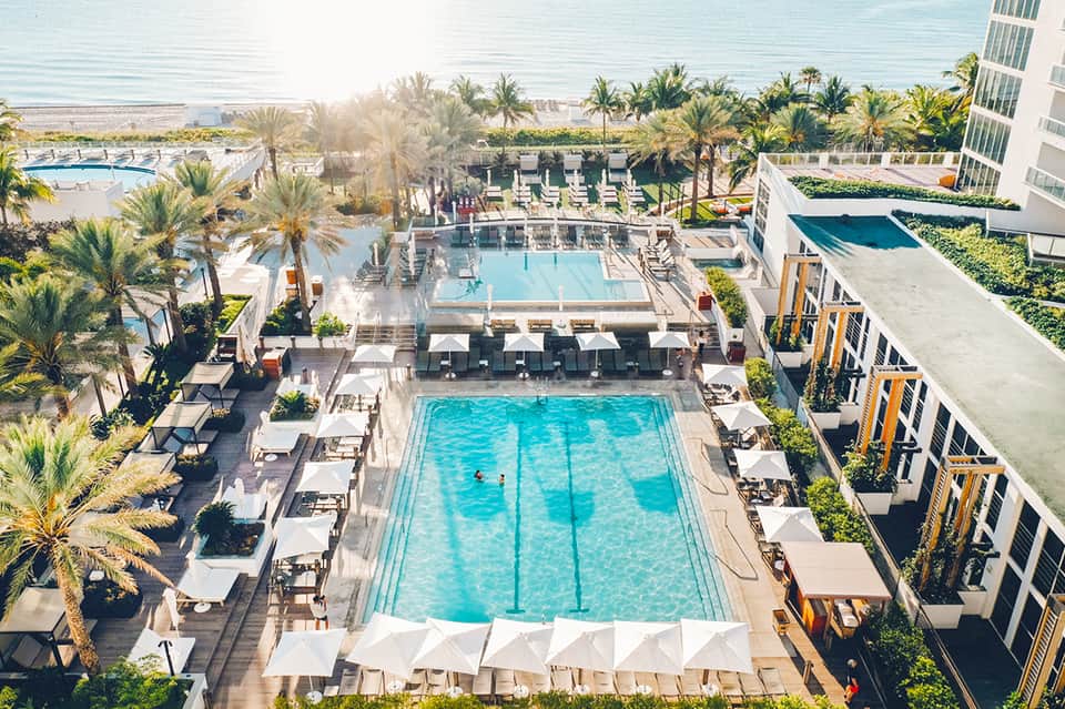 Aerial view of beachfront resort pool complex surrounded by palm trees, loungers, and modern hotel building