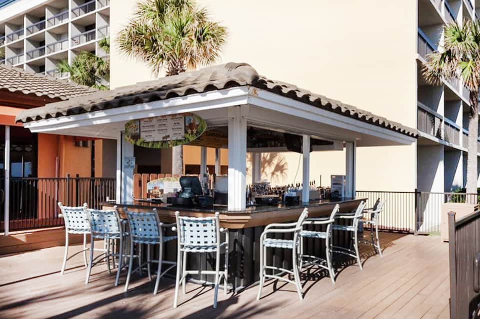 Beachfront bar patio with palm trees, wavy corrugated roof, and high-top seating
