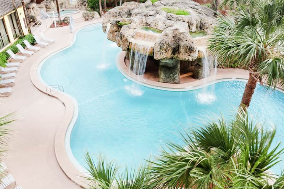 Aerial view of curved turquoise swimming pool with waterfall feature and palm trees surrounding deck