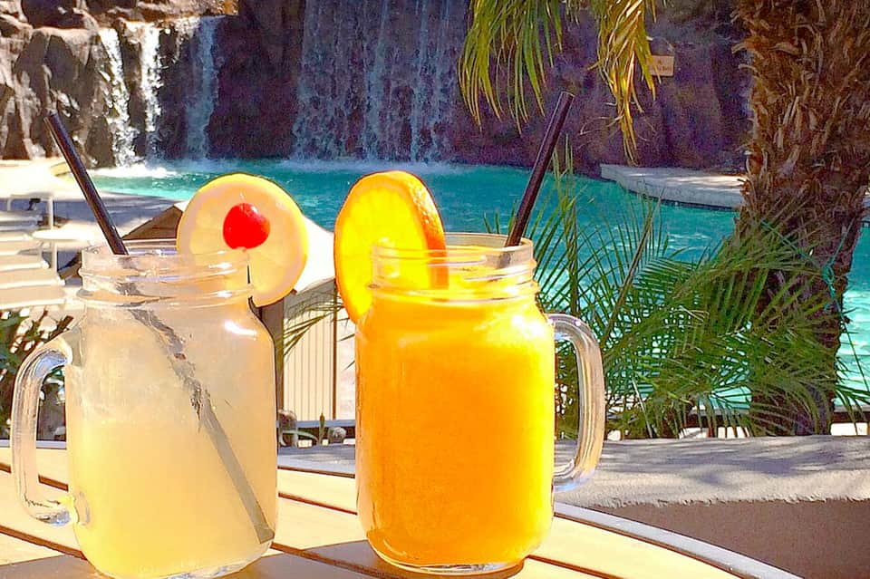 Two colorful tropical cocktails with fruit garnishes on a table overlooking turquoise water and palm trees