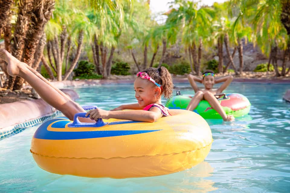 Children playing on colorful inflatable tubes in a tropical resort pool surrounded by palm trees