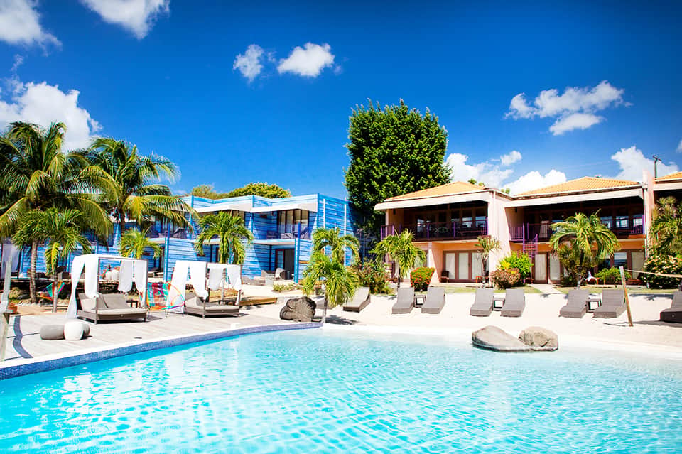 Resort pool with colorful blue and white buildings, palm trees, and lounge chairs under sunny sky
