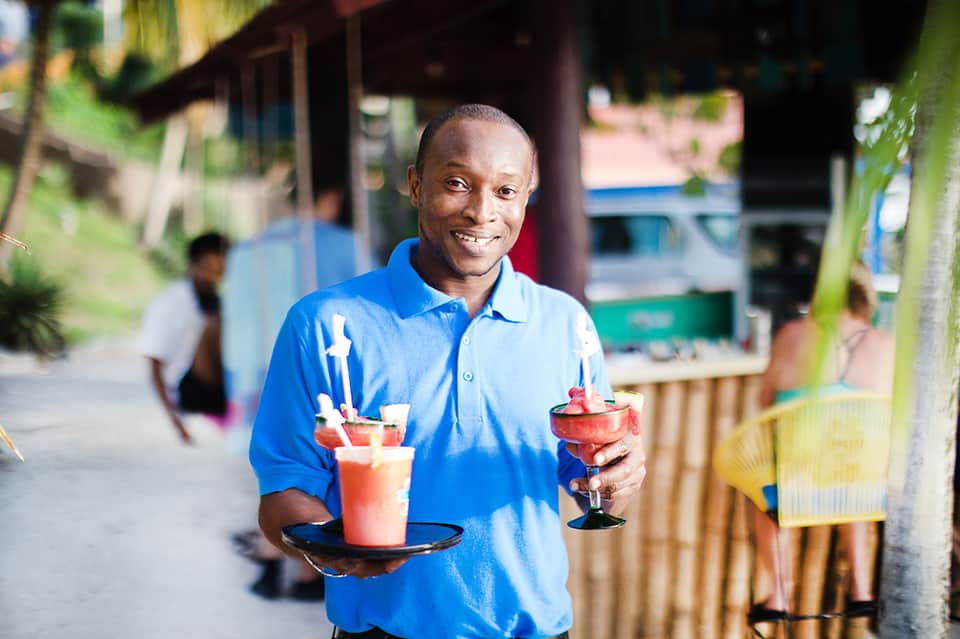 Server in blue shirt smiling while holding tropical beverages on a casual street setting