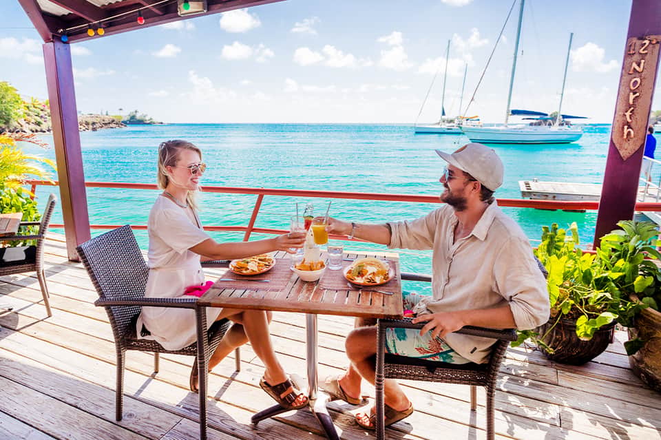 Couple dining on waterfront deck with turquoise ocean, sailboats, and tropical vegetation in background