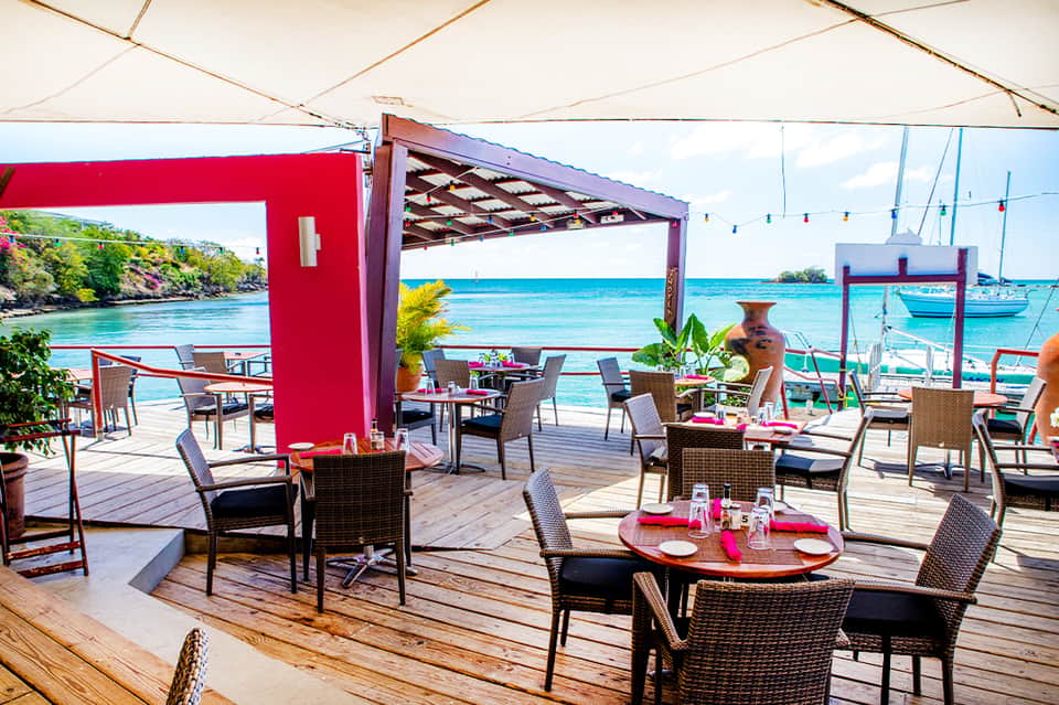 Beachfront dining area with red accents, wicker chairs, wooden deck, and sailboats visible in turquoise water
