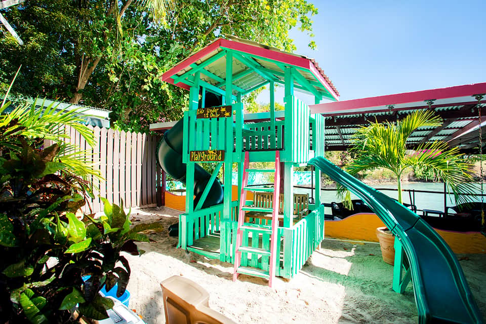 Bright green and red elevated playhouse with slide on sandy beach surrounded by tropical vegetation