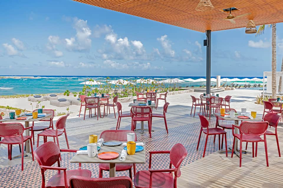 Beachfront dining area with red chairs, patterned floor, wooden shade structure, and ocean views