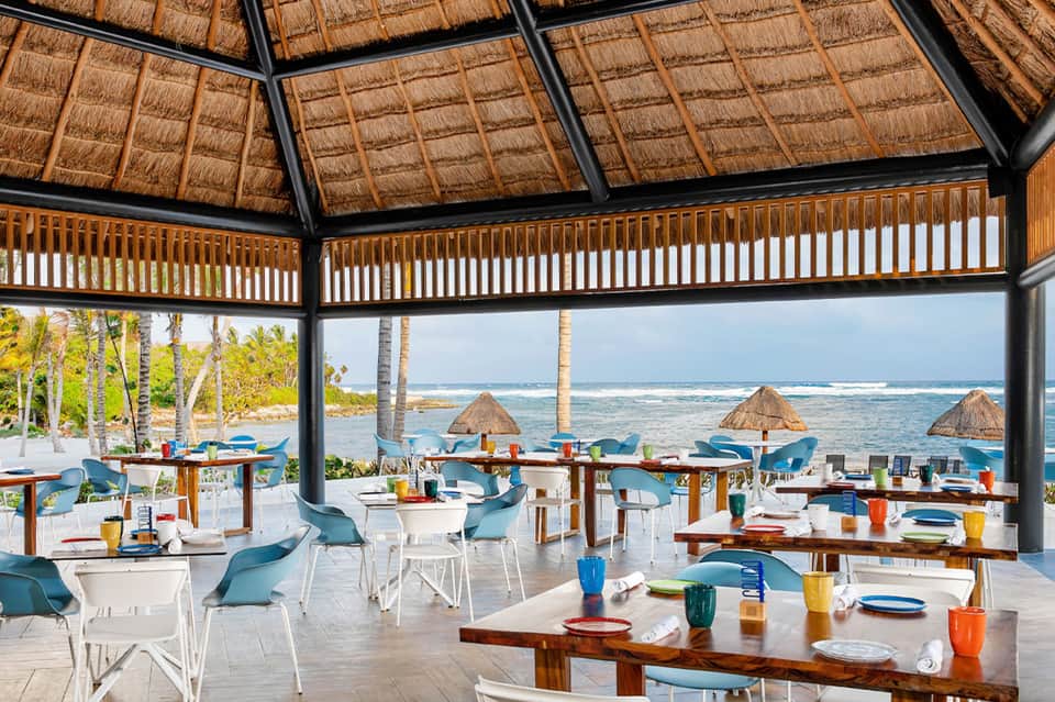 Beachfront dining pavilion with wooden tables, thatched umbrellas, ocean view, and palm trees
