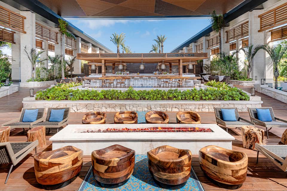 Modern courtyard with fire pit, wooden seating, bar area, and palm trees at midday