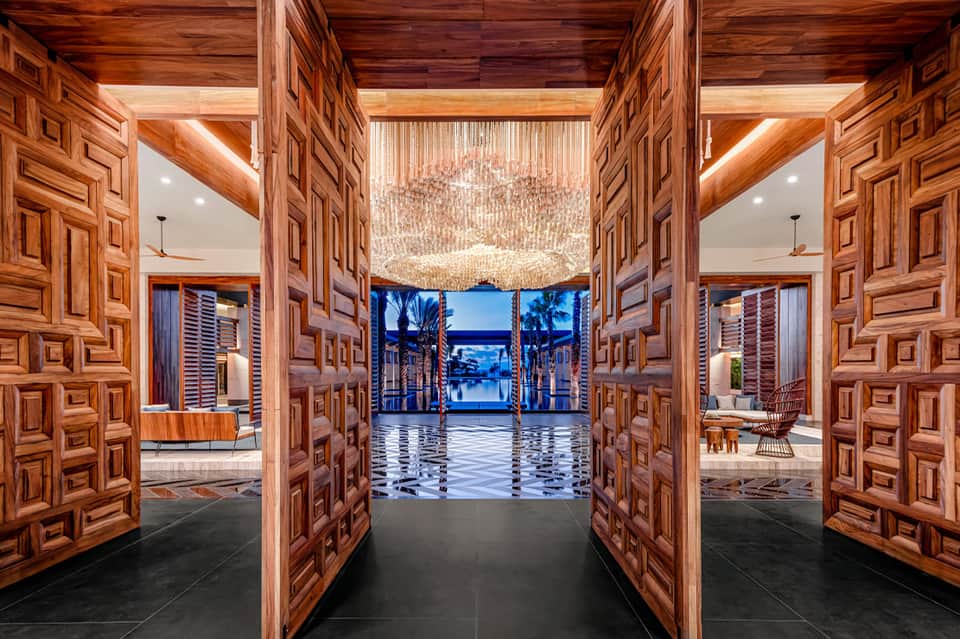 Wooden carved doors opening to ocean view terrace with chandelier and palm trees at dusk
