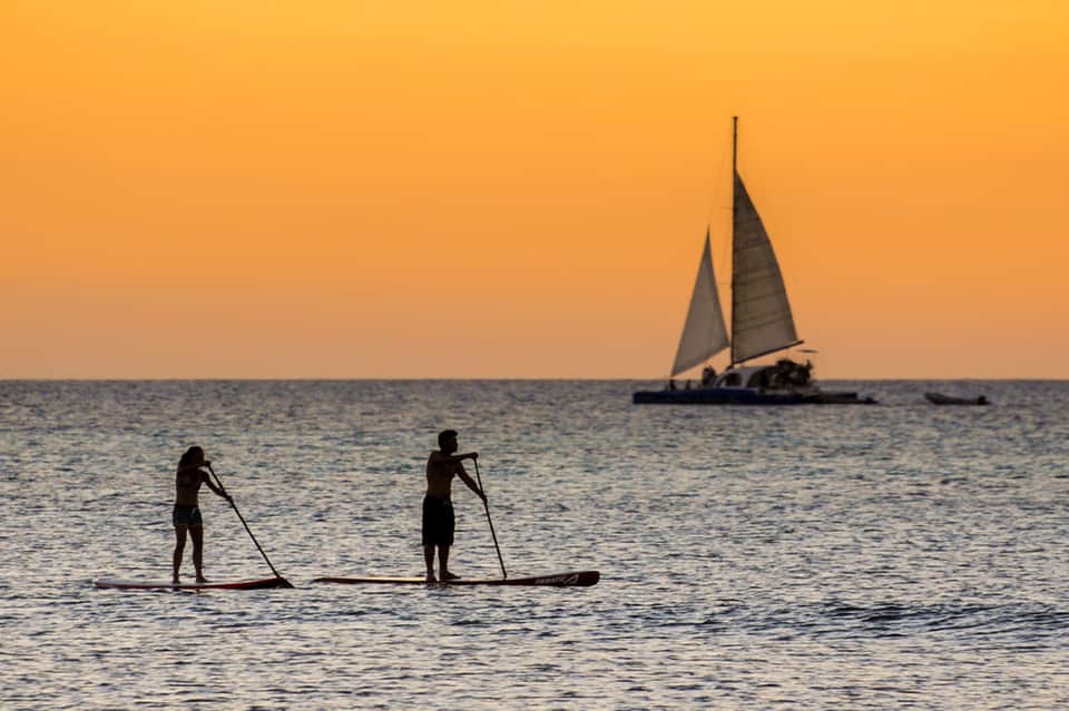 Silhouette of two paddleboarders at sunset with sailboat and orange sky over water