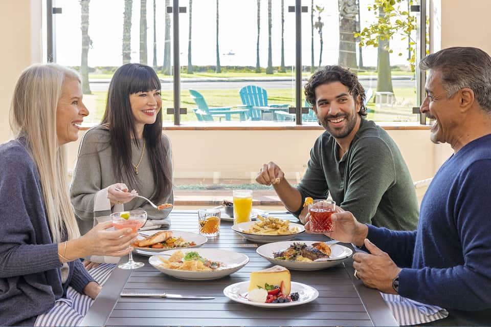 Four adults dining outdoors on waterfront patio with palms visible through windows behind them