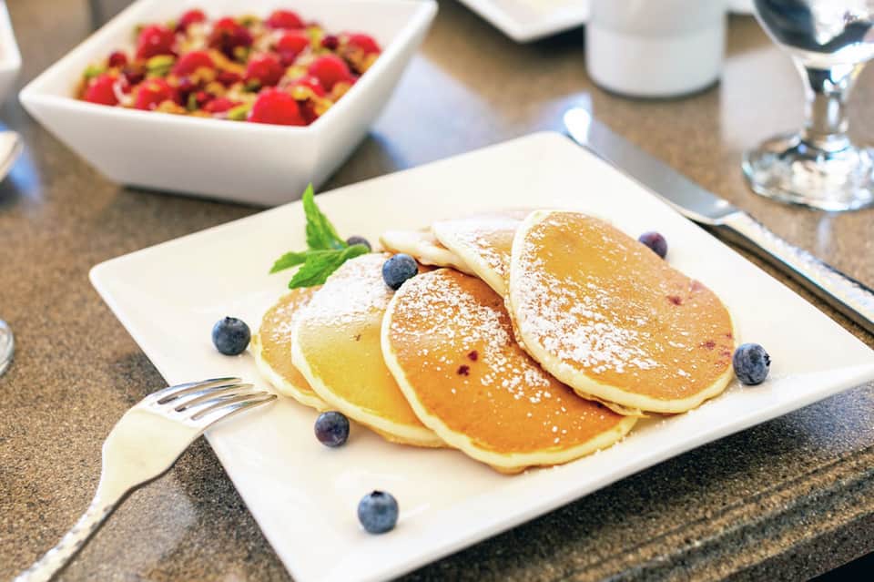 Plate of powdered sugar pancakes with blueberries, fresh berries in bowl, and table settings