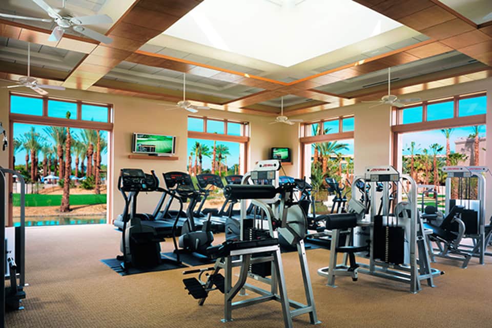 Bright fitness center with cardio equipment, large windows overlooking tropical gardens and palm trees