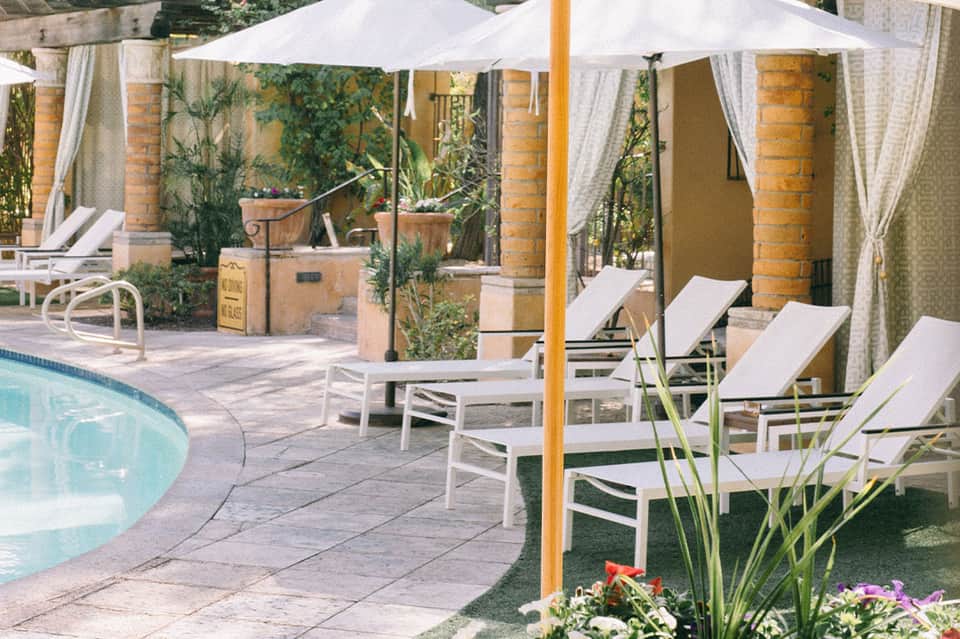Resort pool area with white umbrellas, lounge chairs, brick columns, and lush tropical landscaping