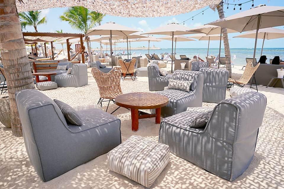Beachfront lounge area with gray upholstered seating, umbrellas, palm trees, and ocean view