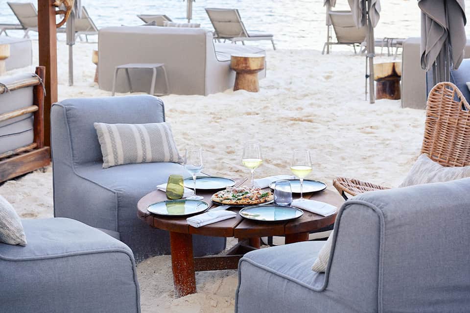 Beachfront dining setup with gray upholstered chairs, round wooden table, and wine glasses