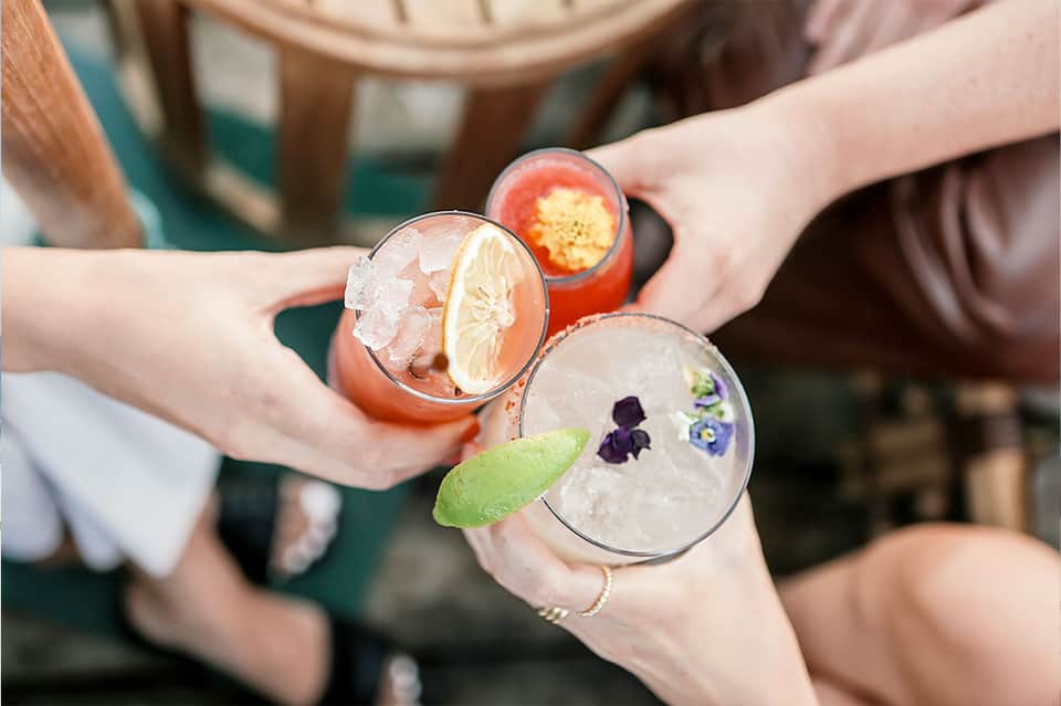 Hands holding three colorful cocktails garnished with citrus and edible flowers